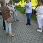 dyrektor czyta ciekawostki historyczne, uczestnicy słuchają stojąc w cieniu na chodniku