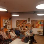 Seniorzy Klubu „Uśmiech Seniora” siedzą na krzesełkach i słuchają o twórczości Alexandra Jolliena