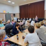 Seniorzy Klubu Seniora siedzą przy stolikach i słuchają poezji Jana Kochanowskiego