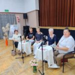 Seniorki siedzą przy stolikach i czytają głośno pieśni, fraszki i treny Jana Kochanowskiego z okazji Narodowego Czytania