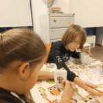 Dziewczynki siedzą przy stole, jedna z nich maluje grzybka