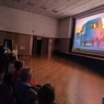 Seniorzy Klubu Seniora „Lokomotywa 60 +” siedzą na krzesełkach i oglądają seans filmowy