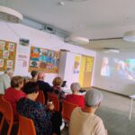 Seniorzy Klubu „Uśmiech Seniora” siedzą na krzesełkach i oglądają polską komedię