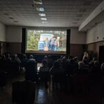 Seniorzy Klubu Seniora „Lokomotywa 60 +” siedzą na krzesełkach i oglądają seans filmowy