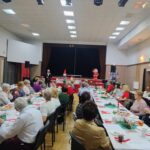 Seniorzy siedzą przy wspólnych stołach i słuchają przemowy dyrektora Miejskiego Ośrodka Pomocy Społecznej Centrum Opieki Socjalnej w Zduńskiej Woli pana Krzysztofa Bejmerta