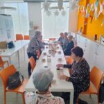 Seniorzy siedzą przy stole podczas słodkiego poczęstunku w postaci pączków, piją herbatę i kawę