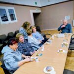 Seniorzy siedzą przy stole podczas słodkiego poczęstunku w postaci pączków, piją herbatę i kawę