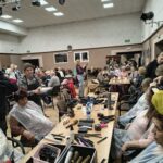 Seniorki siedzą przy stolikach i mają wykonywane zabiegi pielęgnacyjne na twarz