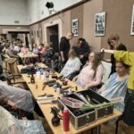 Seniorki siedzą przy stolikach i mają wykonywane zabiegi pielęgnacyjne na włosy