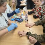 Seniorki siedzą przy stolikach i mają wykonywane zabiegi pielęgnacyjne na włosy